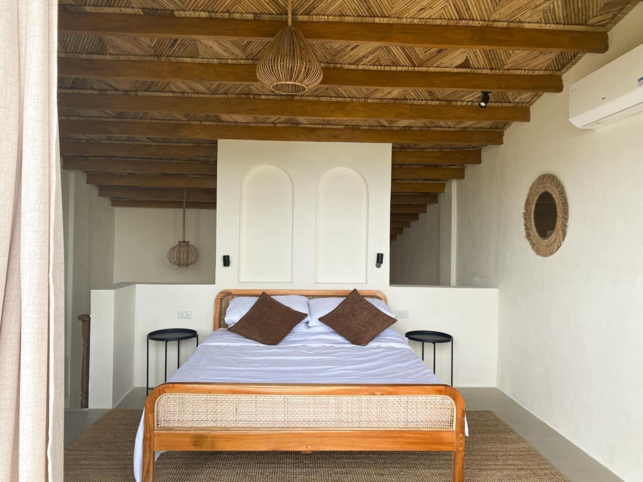 Une chambre à coucher avec un lit en bois, deux tables de chevet, une tête de lit en osier, un ventilateur de plafond et un miroir fixé au mur.