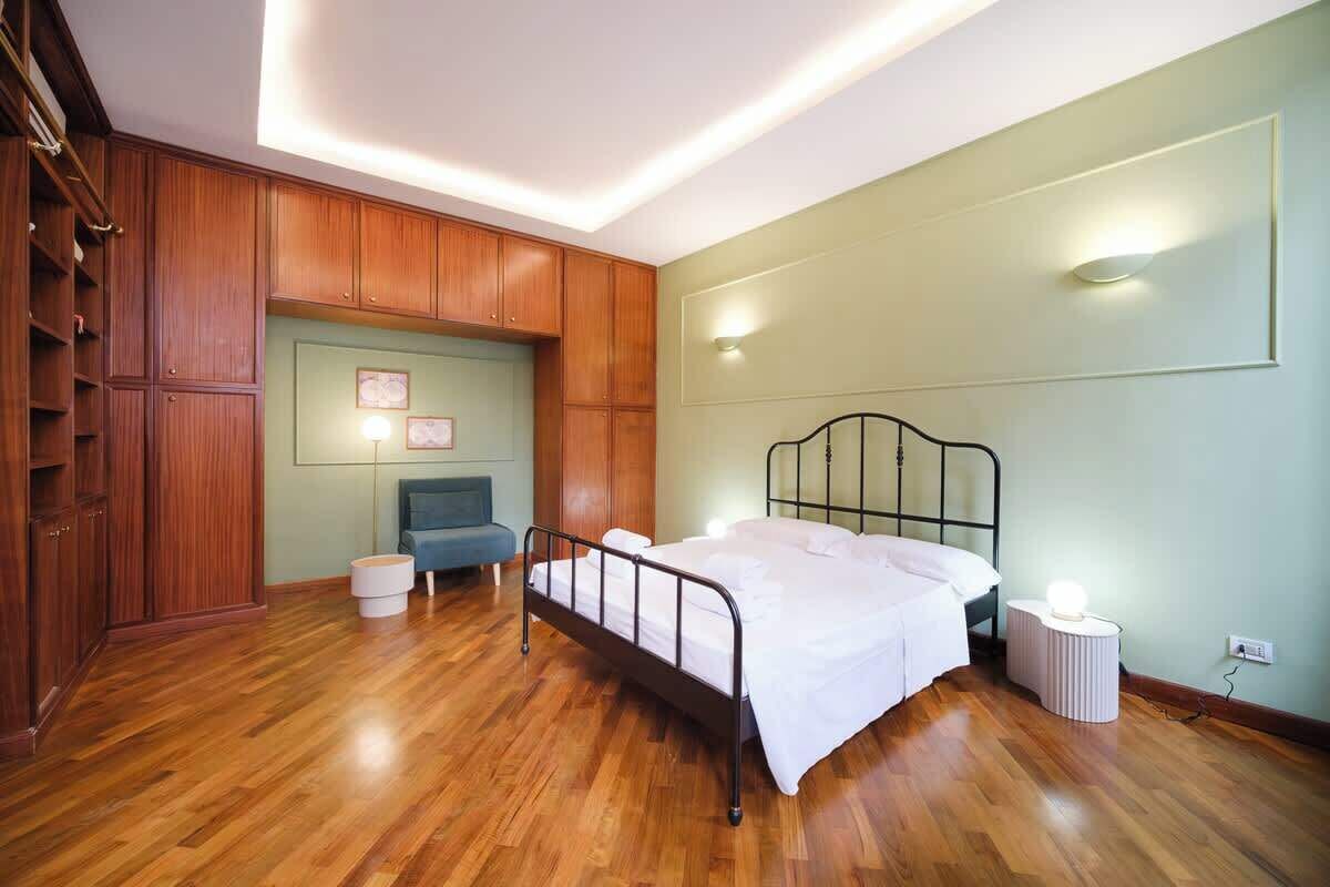 A bedroom with a wooden floor, a metal bed frame, a bedside table with a lamp, and a wardrobe.