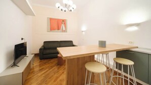 Ein modernes Wohnzimmer mit Holzboden, einer schwarzen Couch, einem Fernseher, einem Holzesstisch mit Hockern und einer Pendelleuchte.