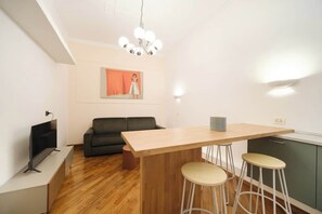 Una sala moderna con piso de madera, un sofá negro, un televisor, una mesa de comedor de madera con taburetes y una lámpara colgante.