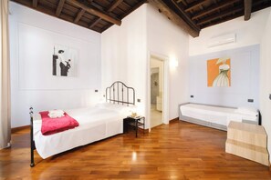 A bedroom with a wooden floor, a bed with a red blanket, a bedside table with a lamp, and a painting on the wall.