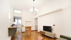 Ein modernes Wohnzimmer mit Holzboden, einem Fernseher, einer Couch, einem Essbereich mit Hocker und einer Küchenzeile.