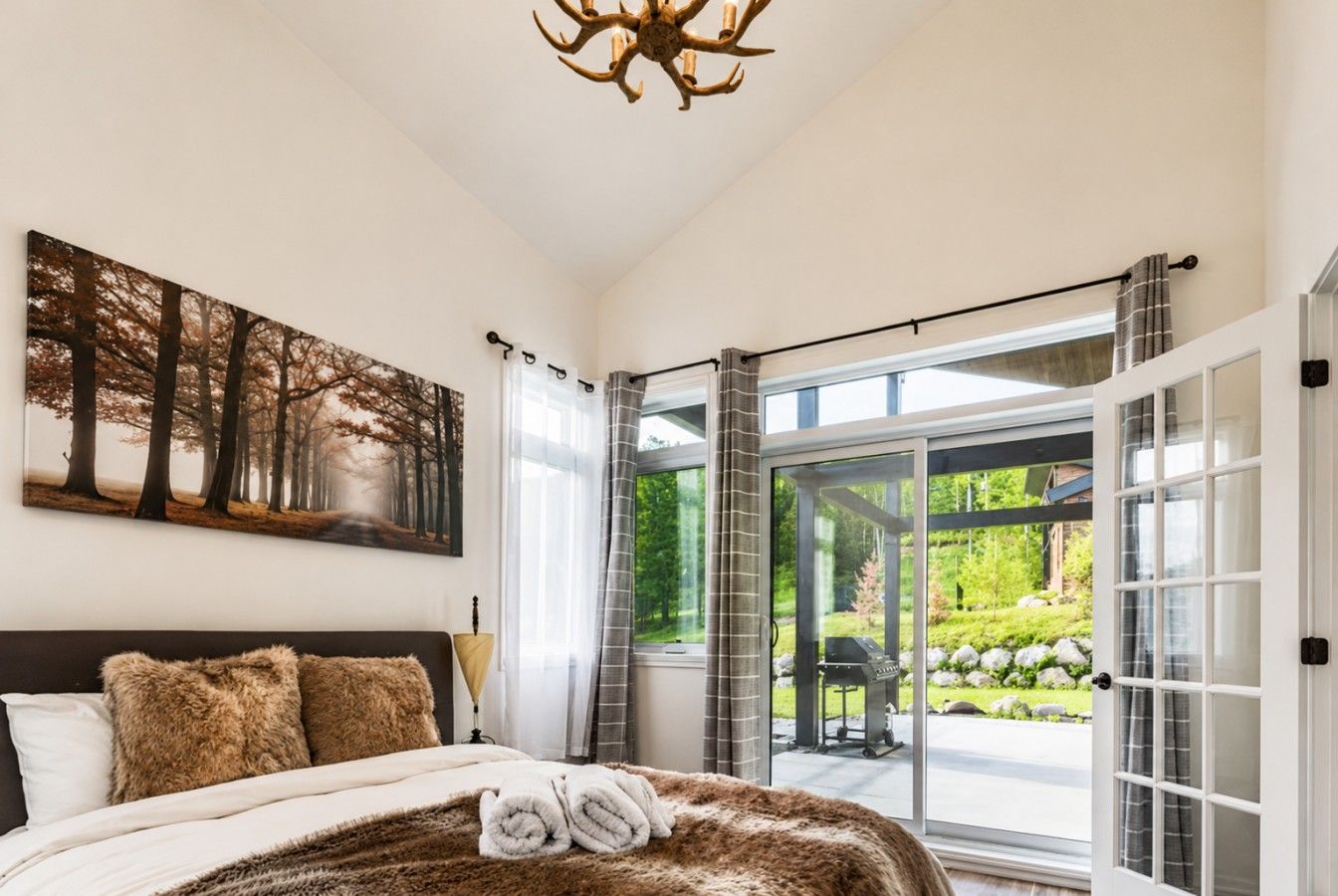 A bedroom with a large bed, a forest-themed painting, and a view of a garden through glass doors.