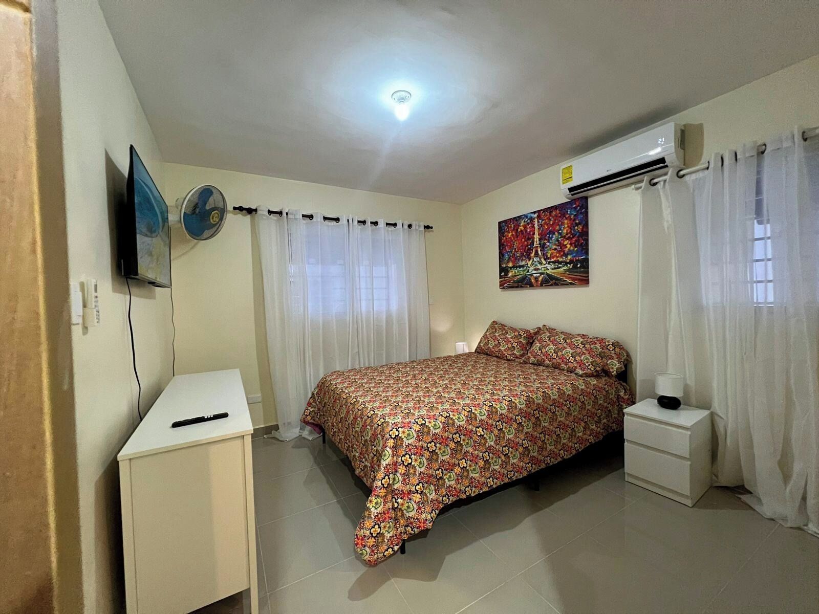 Une chambre à coucher comprenant un lit, un bureau, une lampe sur pied, un climatiseur et un tableau représentant la Tour Eiffel.