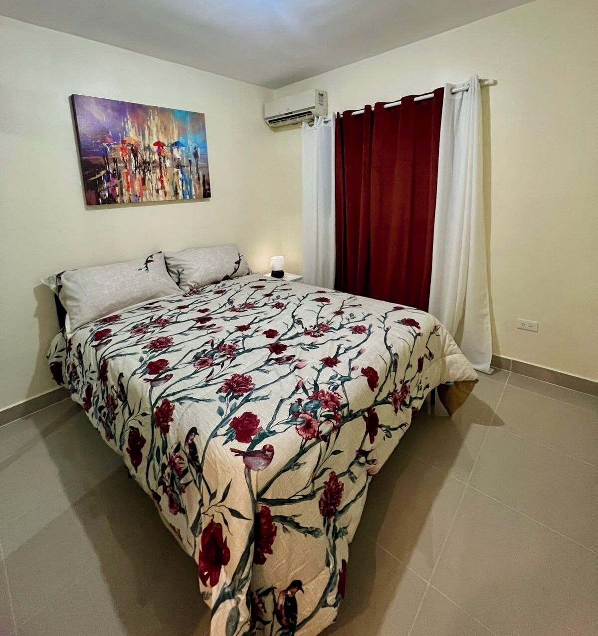 Une chambre avec une couvre-lit à motifs floraux, une table de chevet avec une lampe, un climatiseur fixé au mur et un tableau au-dessus du lit.