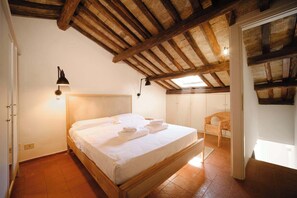 Un dormitorio con techo de madera, cama con ropa blanca, una silla de mimbre y un armario.