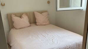 A neatly made bed with a beige headboard and two pillows, a window with blinds, and a light switch on the wall. - Jules' keys (Gruissan)
