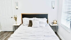 A neatly made bed with white bedding, a dark headboard, and a small bedside table with a lamp and a decorative item. - Downtown - 2 bed, 2 bath (Raleigh)