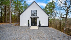 A white building with a triangular roof, a large window, and two small potted plants at the entrance. - Beautiful luxury romantic cabin: hot tub + outdoor swing + secluded+ creek (Broken Bow)