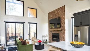 A spacious living area with a green sofa, a fireplace, and a large kitchen with modern appliances. - Beautiful luxury romantic cabin: hot tub + outdoor swing + secluded+ creek (Broken Bow)