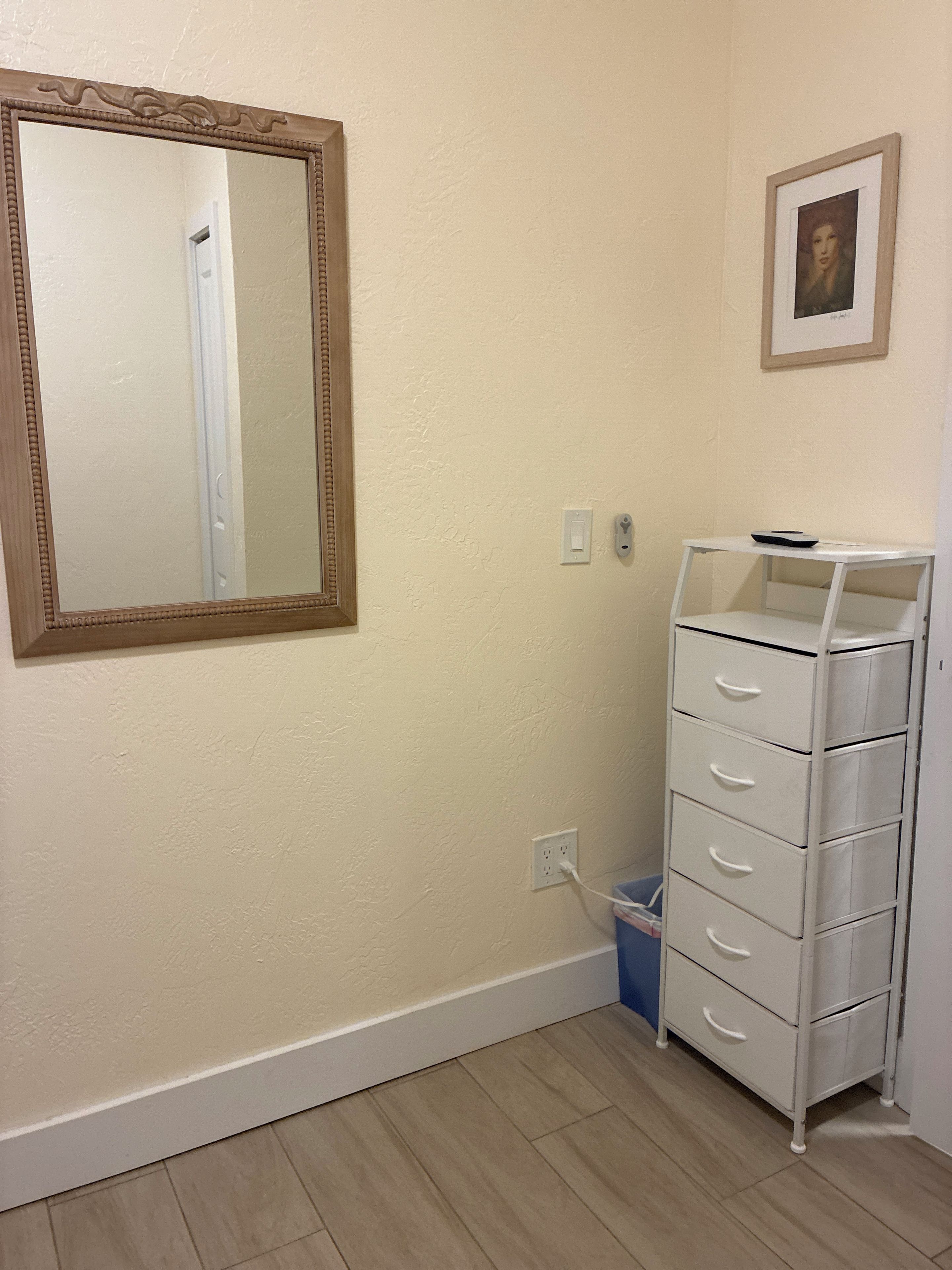 Une pièce avec un grand miroir, une commode blanche à tiroirs, un tableau encadré accroché au mur et un interrupteur.