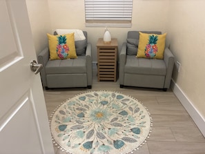 A room with two grey sofas, each with a yellow pillow featuring a pineapple design, a small wooden table with a white lamp, and a round area rug with a floral pattern.