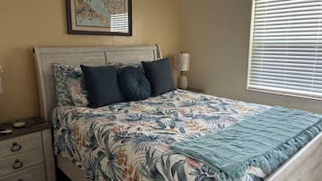 A bedroom with a bed, a nightstand, a lamp, a map on the wall, and a window with blinds.