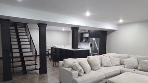 Una sala de estar moderna con un sofá seccional, una cocina con refrigerador de acero inoxidable y una escalera que lleva a un nivel superior.