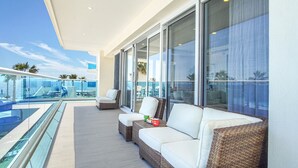 Un balcon avec un canapé blanc, un pouf en osier et une balustrade en verre donnant sur une piscine et des palmiers.