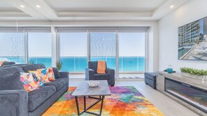 A modern living room with a gray sofa, colorful pillows, a coffee table, and a colorful rug. There is a chair with an orange throw blanket and a large window with a view of the ocean. - Splash Into Summer- Beachfront Condo @ the new Encantame Towers Resort (Puerto Peñasco)