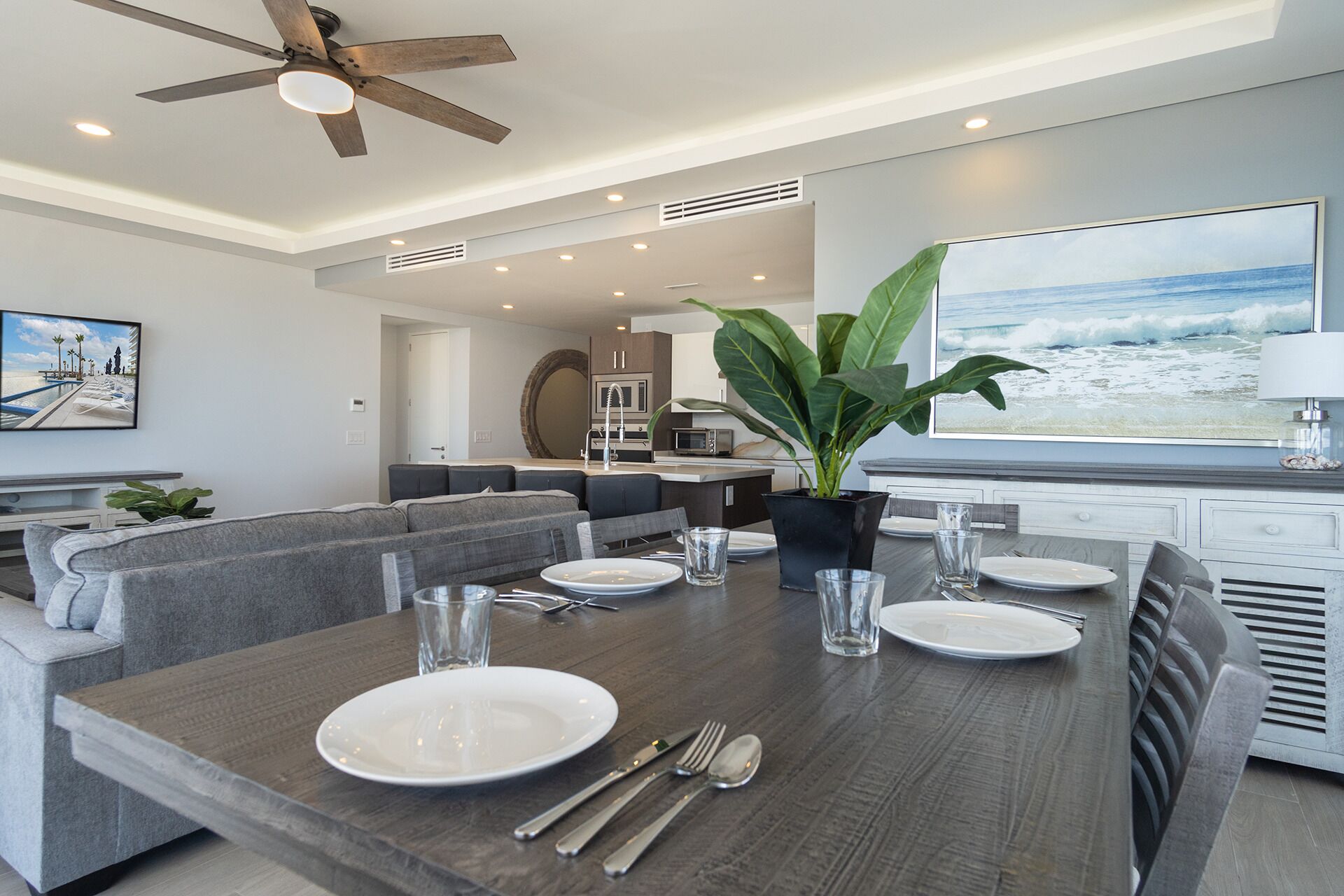 Un espace repas moderne avec une table en bois dressée pour quatre personnes, un canapé gris et une grande fenêtre donnant sur l’océan.