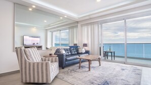 A modern living room with a large sofa, a striped armchair, a coffee table, and a TV on a stand. The room has large windows offering a view of the sea. - Splendid Waterfront 2 Bedroom Velero 1804 Encantame Towers (Puerto Peñasco)