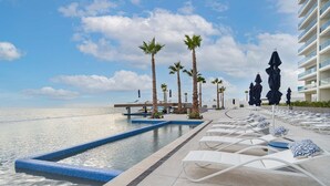 Zona de piscina con sillones de descanso y palmeras, con vistas a una masa de agua.