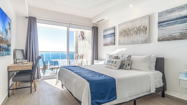 A modern hotel room with a large bed, a desk with a computer, and a balcony with a view of the ocean.