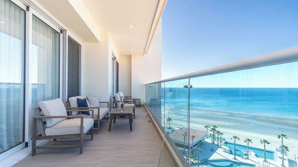 A balcony with a view of the ocean, furnished with chairs and a table, overlooking a pool area with palm trees.