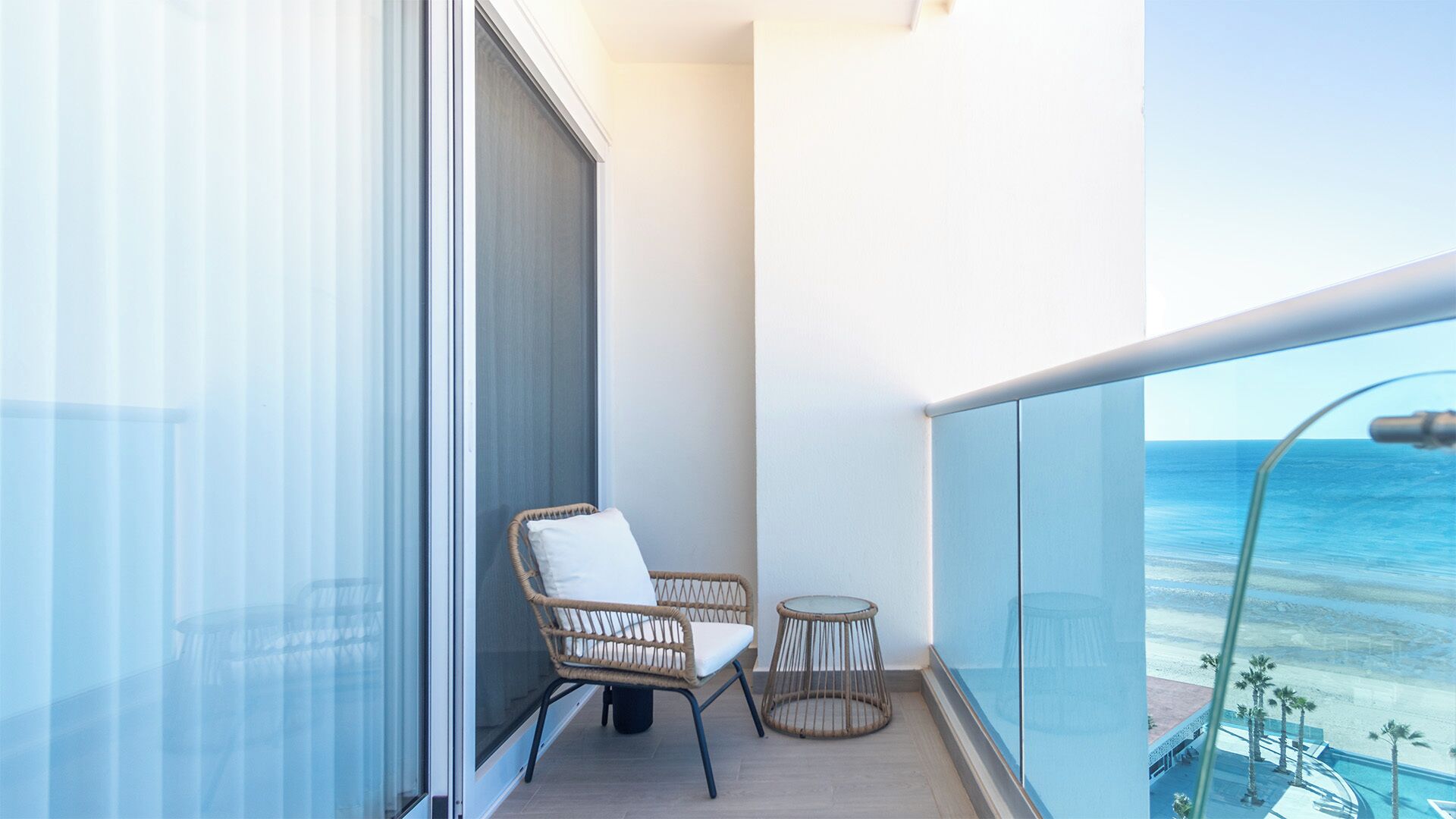 A balcony with a wicker chair and a small table, overlooking a beach.