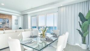 A modern living room with a glass dining table, white furniture, a large window with a view of the ocean, and a balcony with curtains. - Impressive & Beautiful 3 Bedroom Unit Ocean Front Encantame Velero-2301 (Puerto Peñasco)