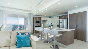 A modern kitchen with a central island, bar stools, and a view of the sea through large windows. - Impressive & Beautiful 3 Bedroom Unit Ocean Front Encantame Velero-2301 (Puerto Peñasco)