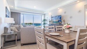 A modern dining area with a wooden table, chairs, and a view of the ocean through large windows. - Encantame Towers Viento 304 (Puerto Peñasco)