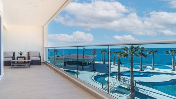 A balcony with a view of a pool and palm trees, a seating area with a sofa and table, and a clear sky. - Encantame Towers Viento 304 (Puerto Peñasco)