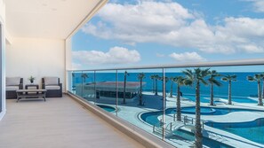 A balcony with a view of a pool and palm trees, a seating area with a sofa and table, and a clear sky. - Encantame Towers Viento 304 (Puerto Peñasco)