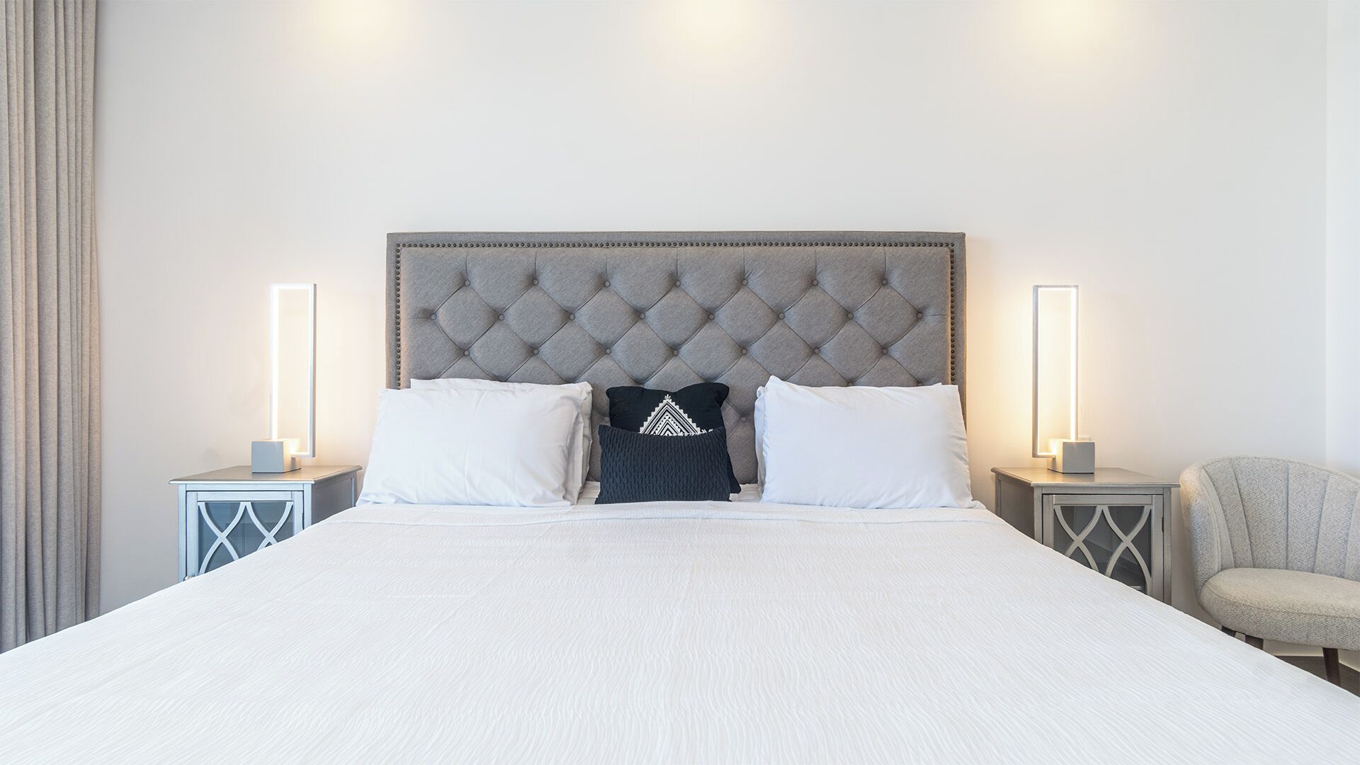 A large bed with a tufted headboard, two bedside tables with lamps, and a chair.