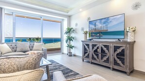 A modern living room with a large window overlooking a beach, a flat-screen TV, and a decorative cabinet. - Encantame Towers Viento 304 (Puerto Peñasco)
