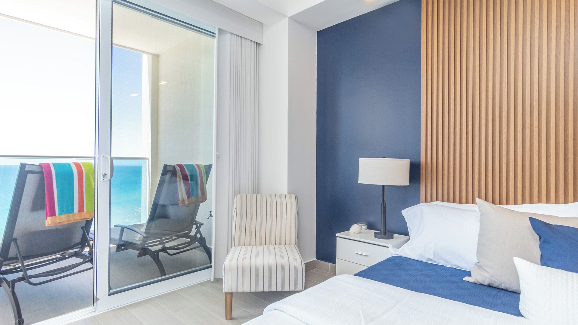 A hotel room with a bed, a chair, a nightstand, a lamp, and a view of the ocean through a sliding door.