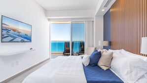 A modern hotel room with a large bed, a TV displaying a beach scene, a balcony with a view of the sea, and a wooden panel wall. - Encantame Towers Viento 1503 (Puerto Peñasco)
