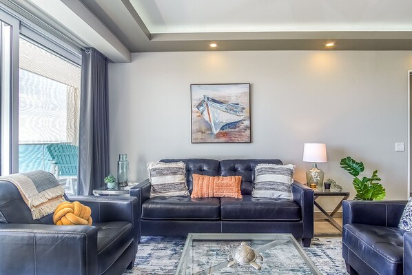 A modern living room with a black leather sofa, a glass coffee table, and a large window with curtains. - Lovely 2 Bedroom Condominium (Puerto Peñasco)