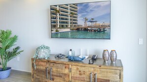 A framed picture of a hotel with a pool and palm trees, a wooden cabinet with decorative items, and a potted plant. - Lovely 2 Bedroom Condominium (Puerto Peñasco)