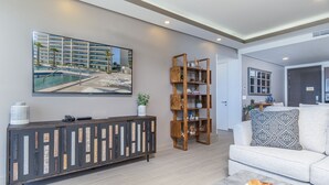 A modern living room with a large flat-screen TV, a wooden bookshelf, a white sofa with patterned cushions, and a small side table. - Perfect Family 2 Bedroom Condo Encantame Viento 504 (Puerto Peñasco)
