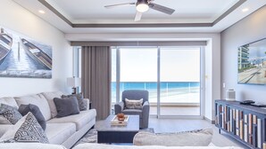 A modern living room with a sofa, armchair, coffee table, and a TV on a cabinet. There is a large window with a view of the beach and ocean. - Perfect Family 2 Bedroom Condo Encantame Viento 504 (Puerto Peñasco)