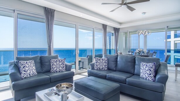 A modern living room with two sofas and a coffee table, large windows with ocean views, and a ceiling fan.