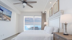 A hotel room with a large bed, a ceiling fan, a nightstand with a lamp, a view of the ocean through a sliding door, and a painting of a beach scene. - Sandy Paws 2 Bedroom 2 Bath Ocean Front Encantame Towers C1604 (Puerto Peñasco)
