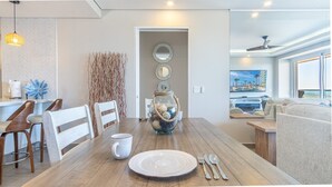 Un área de comedor con una mesa de madera, sillas blancas, un frasco de vidrio decorativo y vista a una piscina y un edificio a través de la ventana.