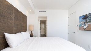 A hotel room with a large bed, a nightstand with a lamp, a door leading to a bathroom, and a wall-mounted TV displaying a beach scene.