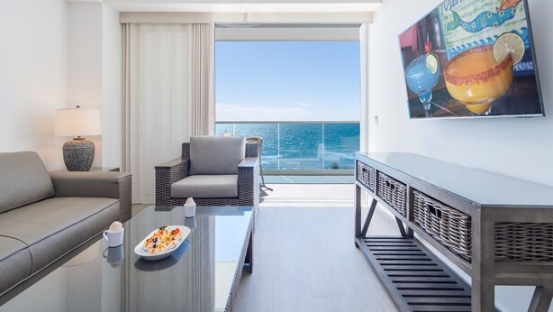 A modern hotel room with a balcony, a sofa, a coffee table with a plate of sushi, and a wall-mounted TV displaying a mermaid-themed artwork.