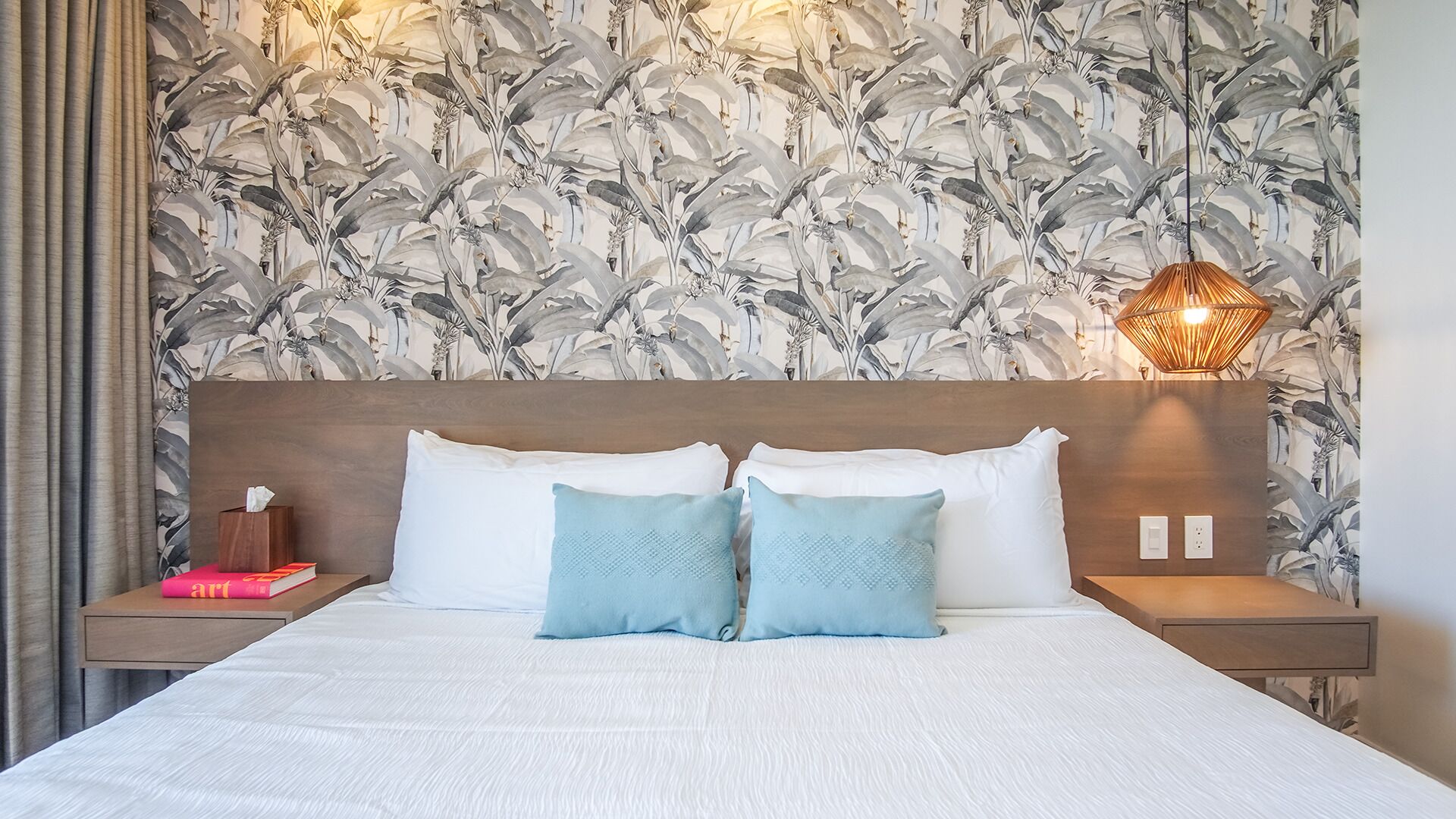 Una habitación de hotel con una cama grande, dos almohadas, una mesita de noche con un libro, un adorno en la pared y una lámpara de pared.