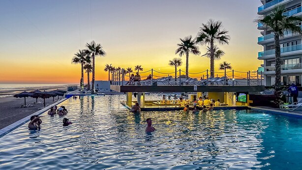 Un área de piscina con vista al atardecer, palmeras y un puente que conecta con un edificio.