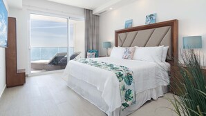 A hotel room with a large bed, a balcony with a view of the sea, and two wall-mounted lamps.