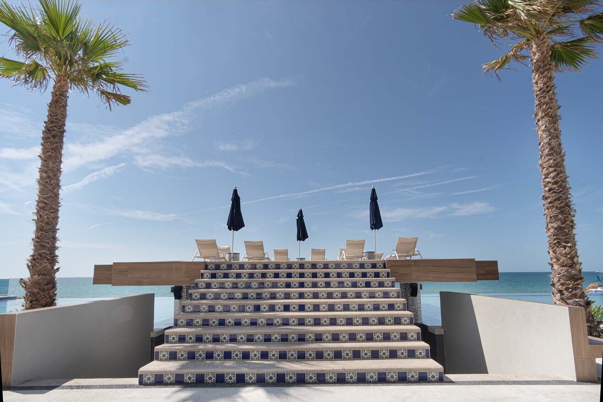 A staircase leading to a beach area with lounge chairs and umbrellas, palm trees, and a clear view of the ocean.