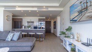 A modern living room with a grey sofa, a coffee table, and a dining area with a bar. There is a kitchen in the background with modern appliances and a large window. - Spectacular 2 Bedroom Ground Floor Condominium Encantame E 105 (Puerto Peñasco)