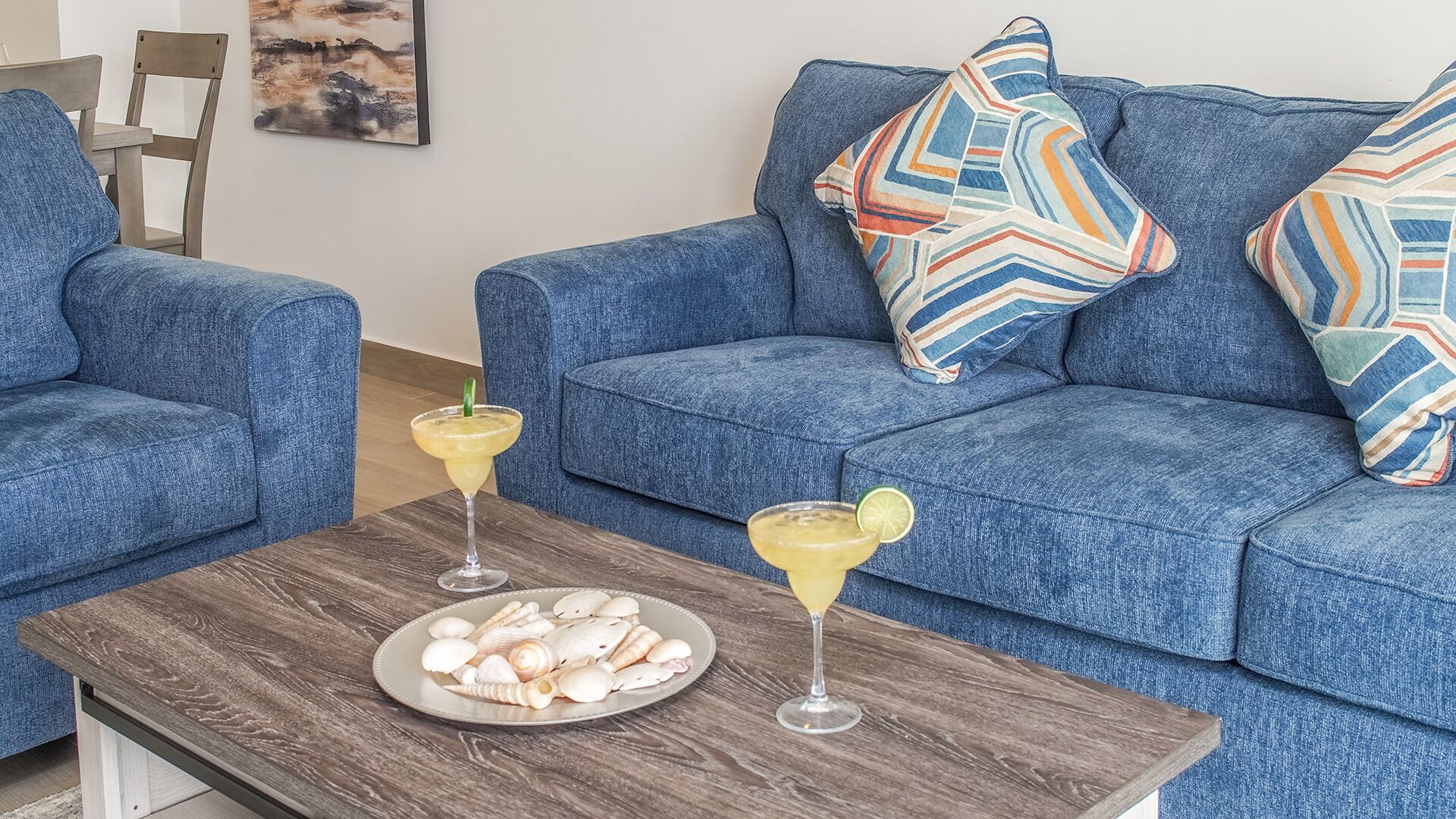 Un salon avec un canapé bleu, deux coussins à motifs, une table basse en bois sur laquelle se trouve une assiette remplie de coquillages et deux boissons.